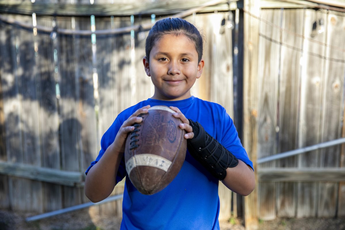A young boy stands in front of a fence holding a football in her hands.