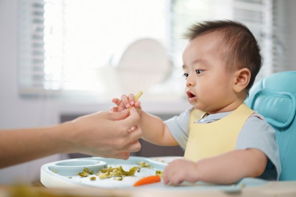 A baby in a high chair that has broccoli and a carrot on the tray. A woman’s hand can be seen handing food to the baby, who is wearing a bib.