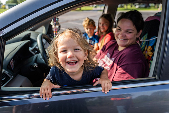 A family sitting in a parked car. One kid leans out through the window toward the camera with a big smile.