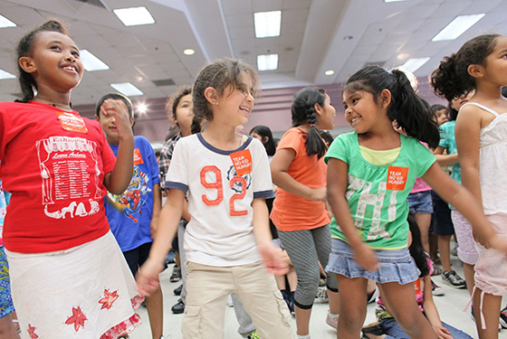 A group of elementary school aged kids dancing and smiling together. Many have No Kid Hungry stickers on their shirts.