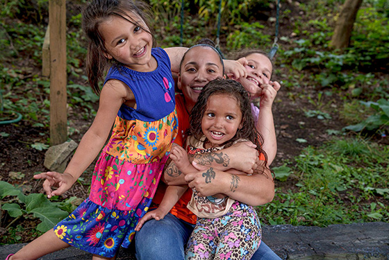 A mom sitting surrounded by her three daughters who are all smiling and making silly faces.