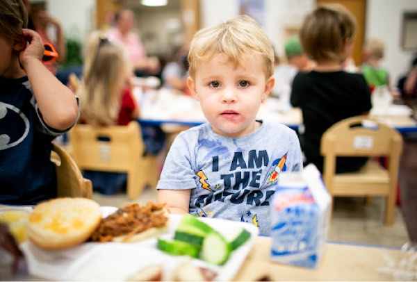 A young boy sitting in a crowded cafeteria. A tray of food is in the forefront. The boy is wearing a shirt that says, “I am the future.