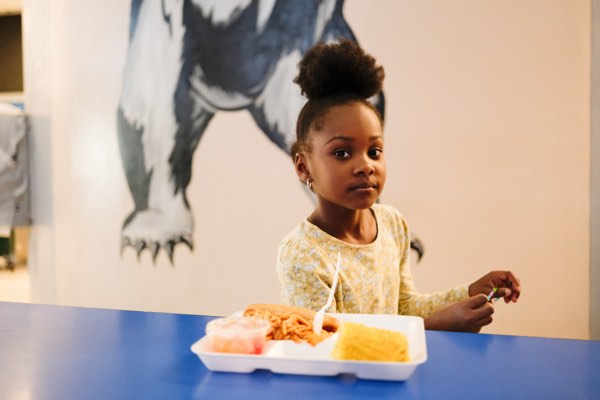 A young girl sits at a lunchroom table in front of a tray of food.